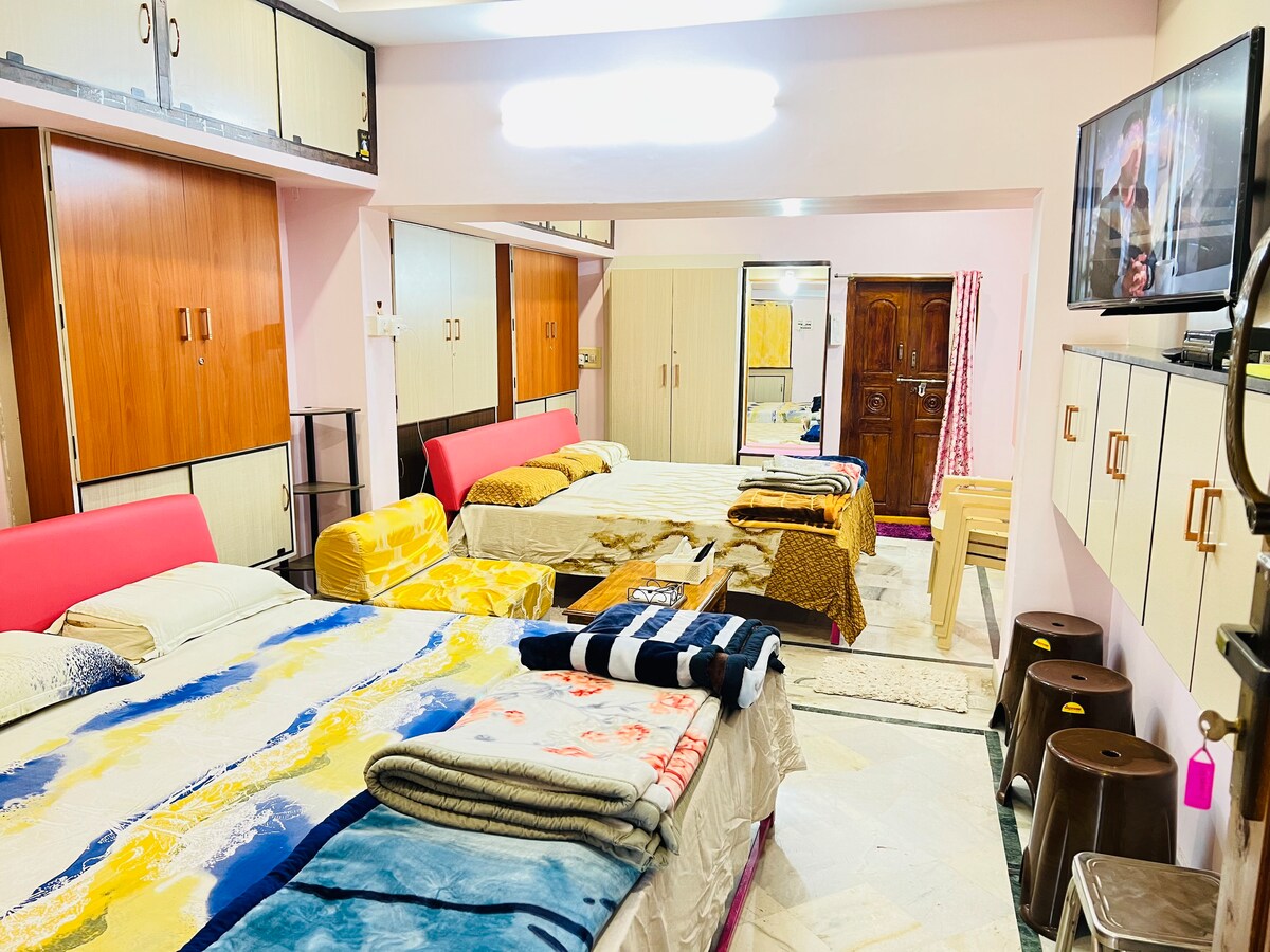 A spacious room features several beds with layered blankets and bedding in various colors. Storage cabinets line the walls, and a wall-mounted television is visible. Light reflects off the marble floor, creating an open and airy atmosphere.