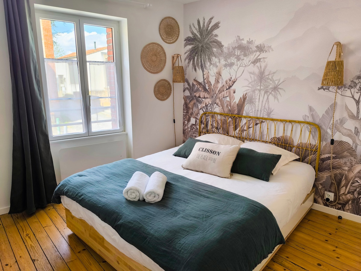 A bright bedroom features a spacious bed adorned with white linens and a dark green throw. Decorative elements include wall art depicting tropical foliage, round woven wall hangings, and two bedside baskets. Natural light enters through a large window, complemented by dark curtains.