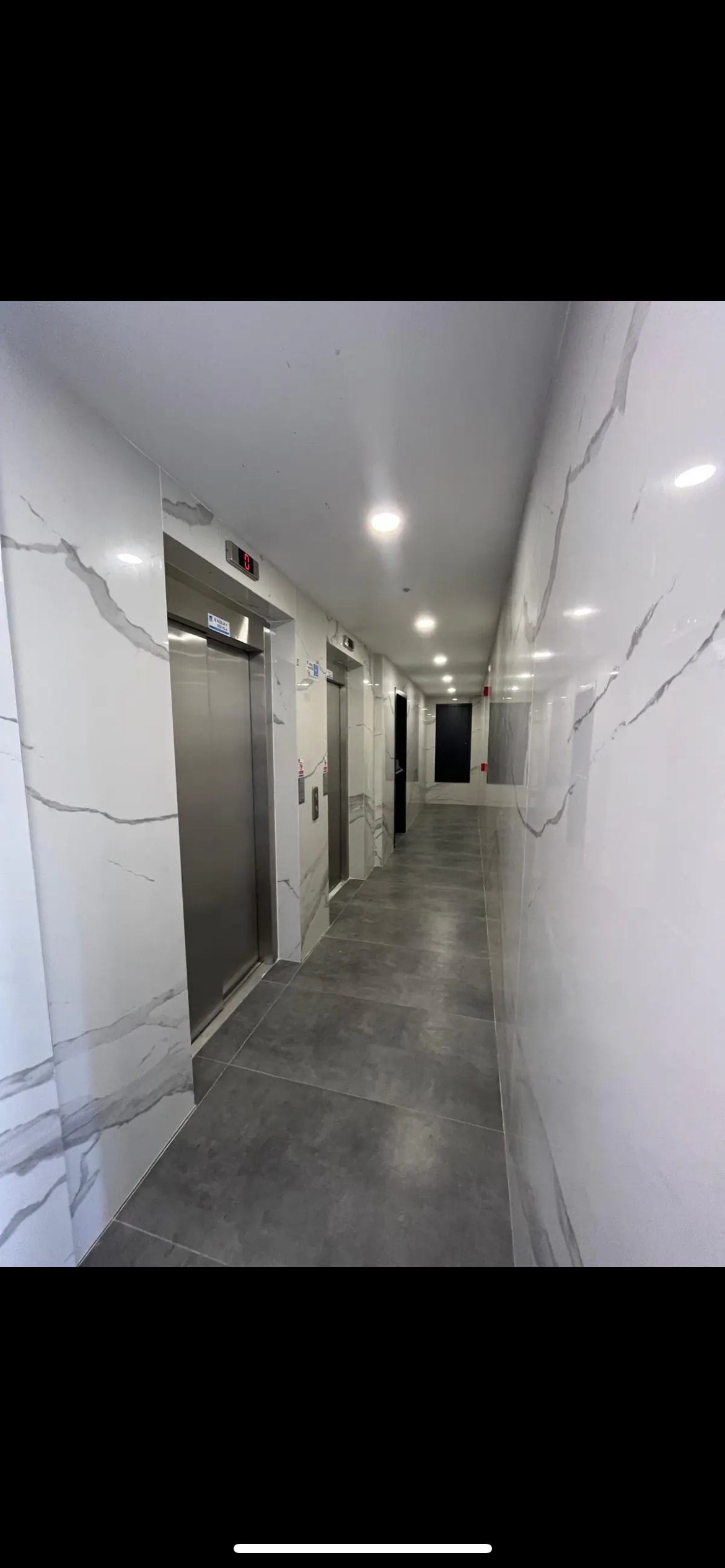 A sleek corridor features modern elevators on one side, complemented by light-colored marble walls. The flooring consists of grey tiles, and a soft lighting enhances the clean and minimalist design.
