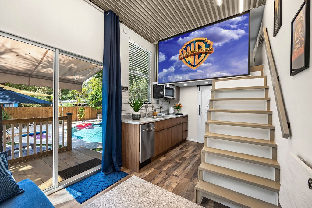 An open kitchen area is seen featuring modern cabinetry and appliances, with a large screen displaying a logo on the wall. Stairs lead to the upper level, while a view of the private pool and lounge area is visible through glass doors.