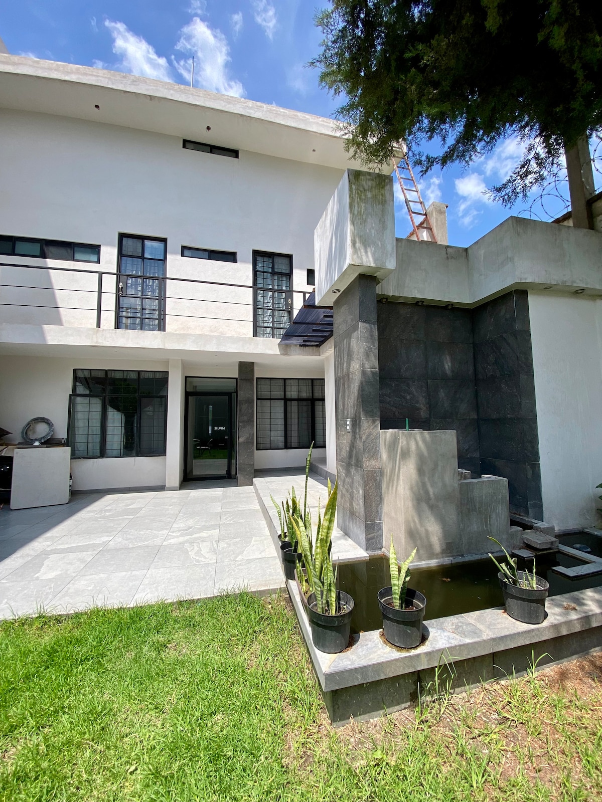 A modern two-story building is highlighted, featuring large windows and a sleek stone exterior. A small water feature is visible, surrounded by potted plants on a grassy area. The sky above is clear, indicating a bright, sunny day.