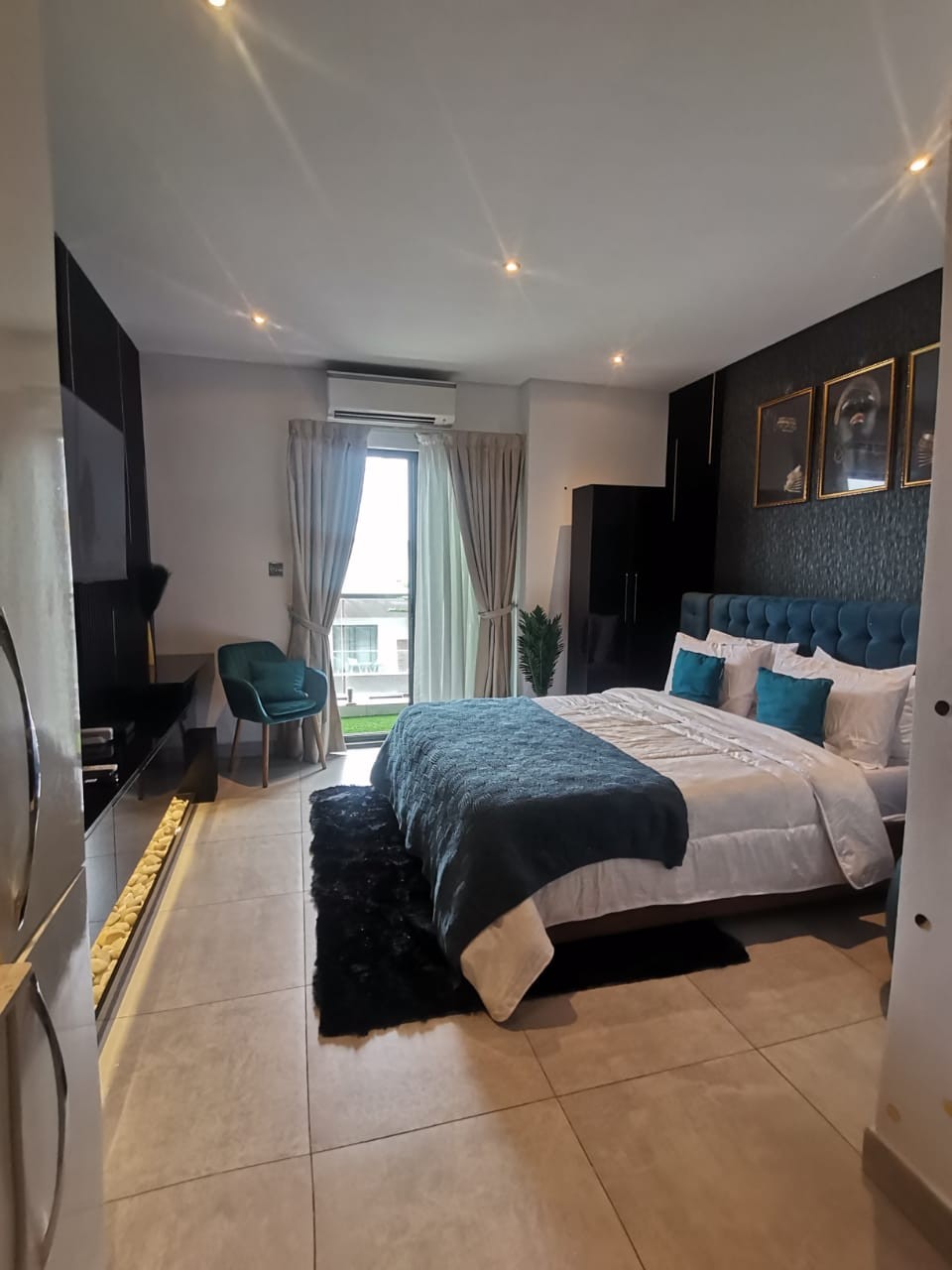 A well-lit bedroom features a spacious bed adorned with a plush blue throw. Stylish dark furnishings and decorative wall art complement the space. Large windows offer a view and natural light, while a green chair adds a touch of color to the room.