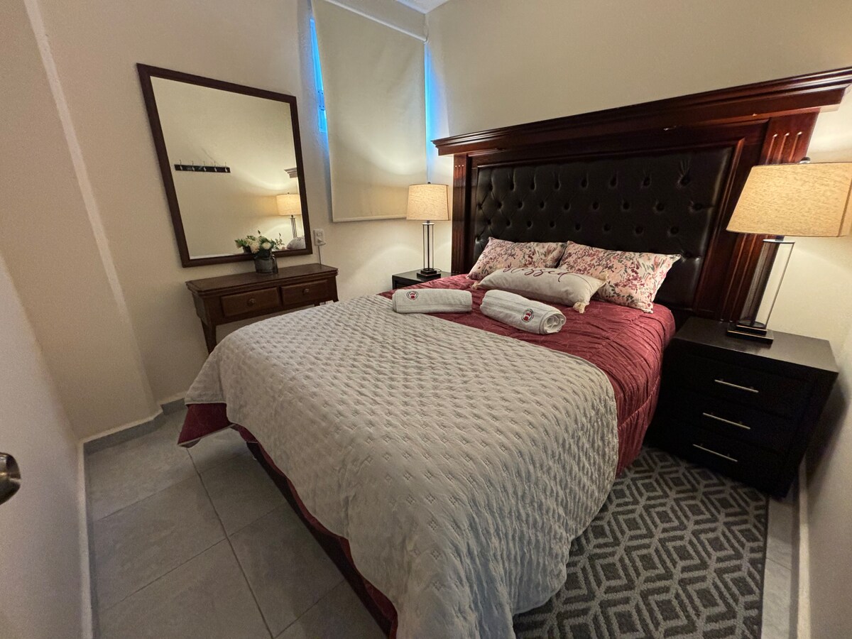 A comfortable bedroom features a spacious bed with decorative pillows and plush bedding. Nightstands with lamps are positioned on either side, illuminating the area. A large mirror reflects the space, and a gentle glow enters through the window with soft treatments.