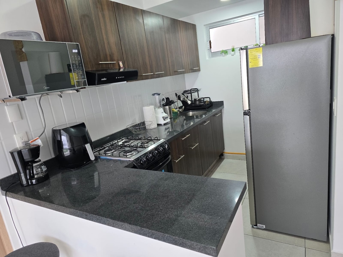 A modern kitchen features dark wood cabinets and a sleek countertop. Basic appliances include a microwave, coffee maker, and kettle. A stainless steel refrigerator stands beside a gas stove, with ample natural light coming from a small window above the sink.