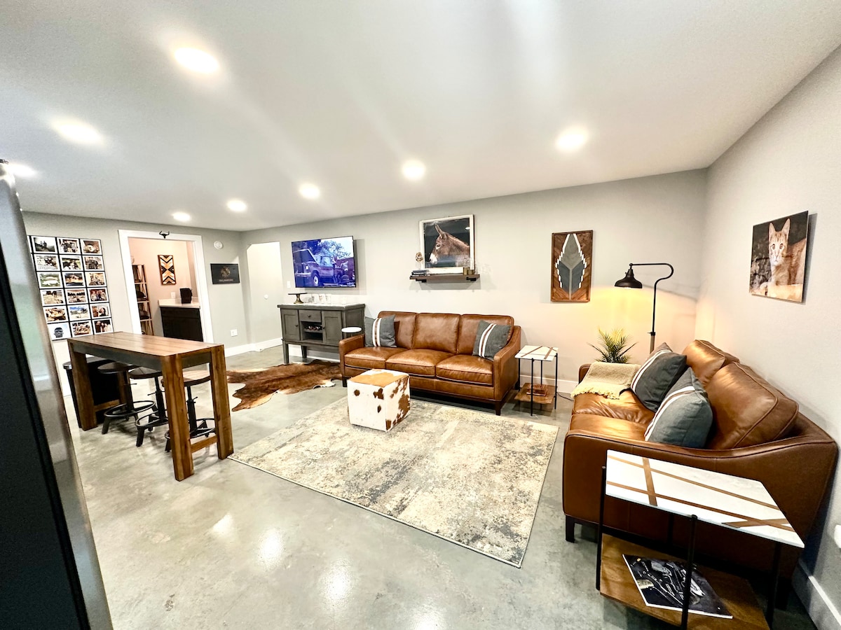 A modern living area is depicted, featuring two leather sofas and a wooden coffee table situated on a textured area rug. A vintage-style TV is mounted on the wall, while bright recessed lighting enhances the spacious feel. A small dining table is positioned in the corner.
