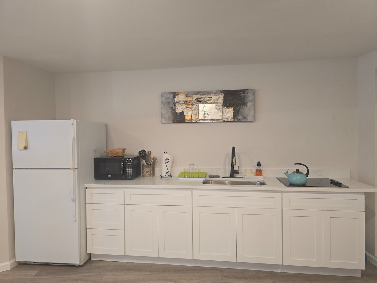 A functional kitchen area showcases modern white cabinetry, featuring a countertop with a sink and stovetop. Essential appliances, including a refrigerator and microwave, are clearly visible. Decorative elements such as a wall-mounted art piece add a touch of style above the countertop.