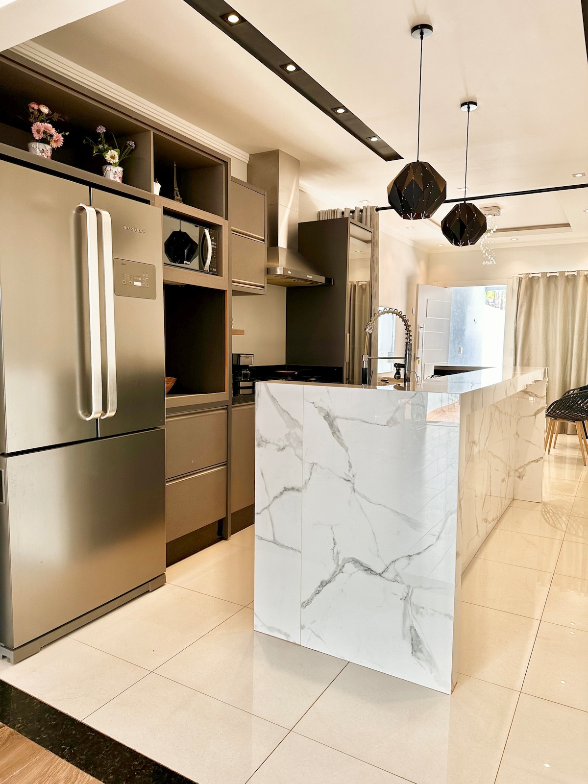 A modern kitchen features sleek cabinetry and a white marble island, enhancing the space. A stainless steel refrigerator stands beside built-in appliances, while pendant lights hang above. Light-colored tiles contribute to a bright, spacious atmosphere, with a window allowing natural light to enter.