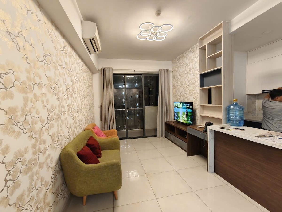 A comfortable living area is presented, featuring a green sofa and a yellow sofa adorned with red cushions. Large glass doors provide access to a balcony, while a television is mounted on the wall opposite. The kitchen area is partially visible, showcasing modern cabinetry.
