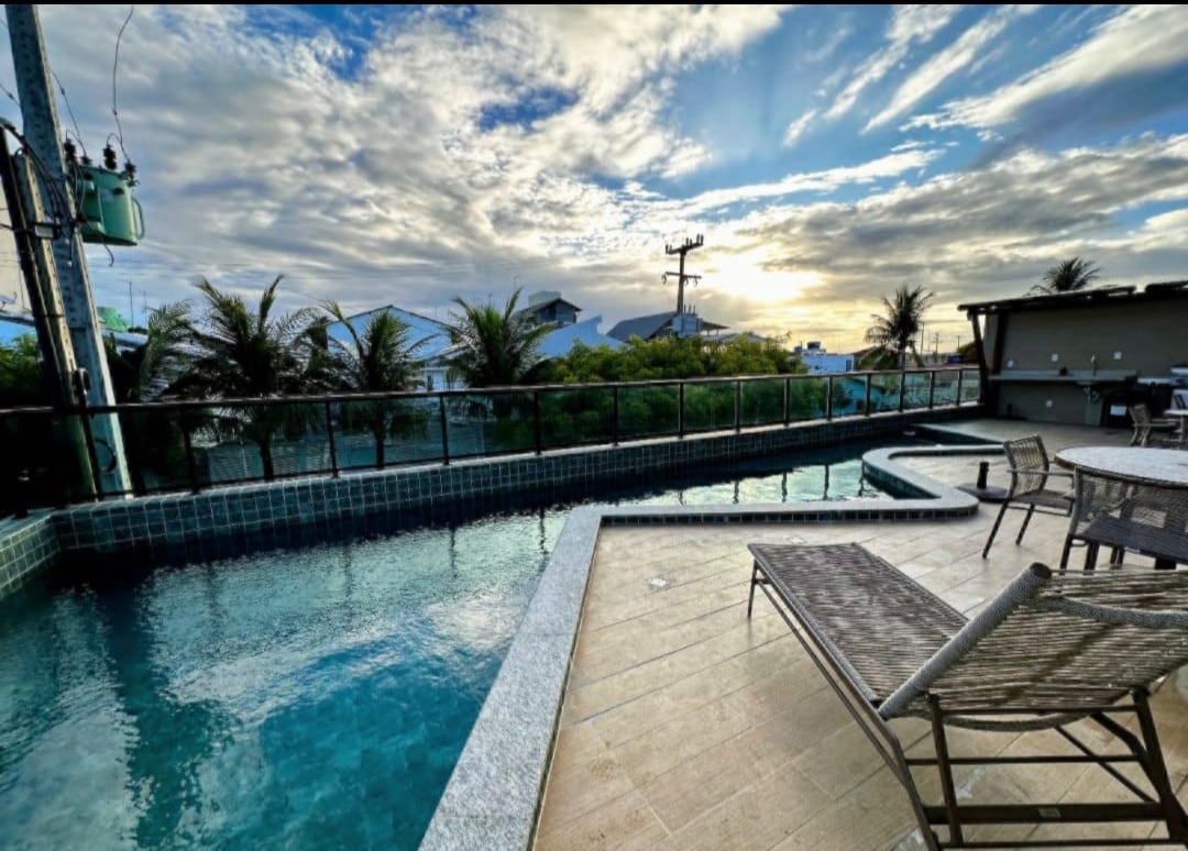 An outdoor area features a swimming pool with a section for relaxation, surrounded by a deck and lounge chairs. Lush greenery and palm trees are visible in the background, complemented by a dramatic sky with clouds and soft sunlight, enhancing the tranquil atmosphere.