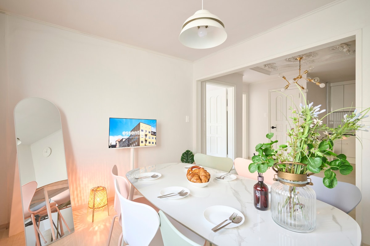 A modern dining area is highlighted by a spacious table set for six, featuring elegant dishware and a decorative vase with fresh greenery. A large mirror reflects light, and a flat-screen TV is mounted on the wall, contributing to a bright, welcoming atmosphere.