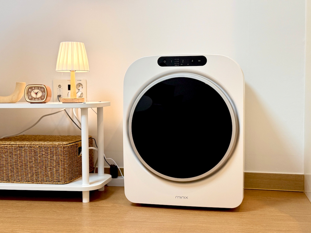 An air purifier is positioned in a well-lit corner of the room. Its modern design features a large circular intake vent. Nearby, a small white lamp and a wooden clock rest on a side table, providing a sense of functionality and comfort to the space.