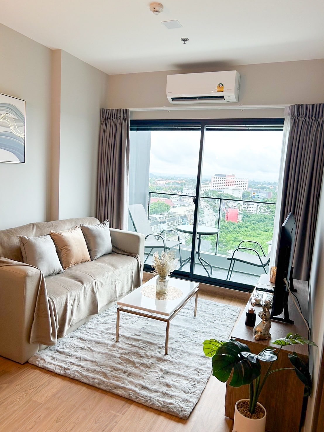 A stylish living area features a comfortable sofa adorned with cushions, a glass coffee table, and a decorative rug. Large windows provide ample natural light and offer views of the surrounding cityscape. A small plant adds a touch of greenery to the space.
