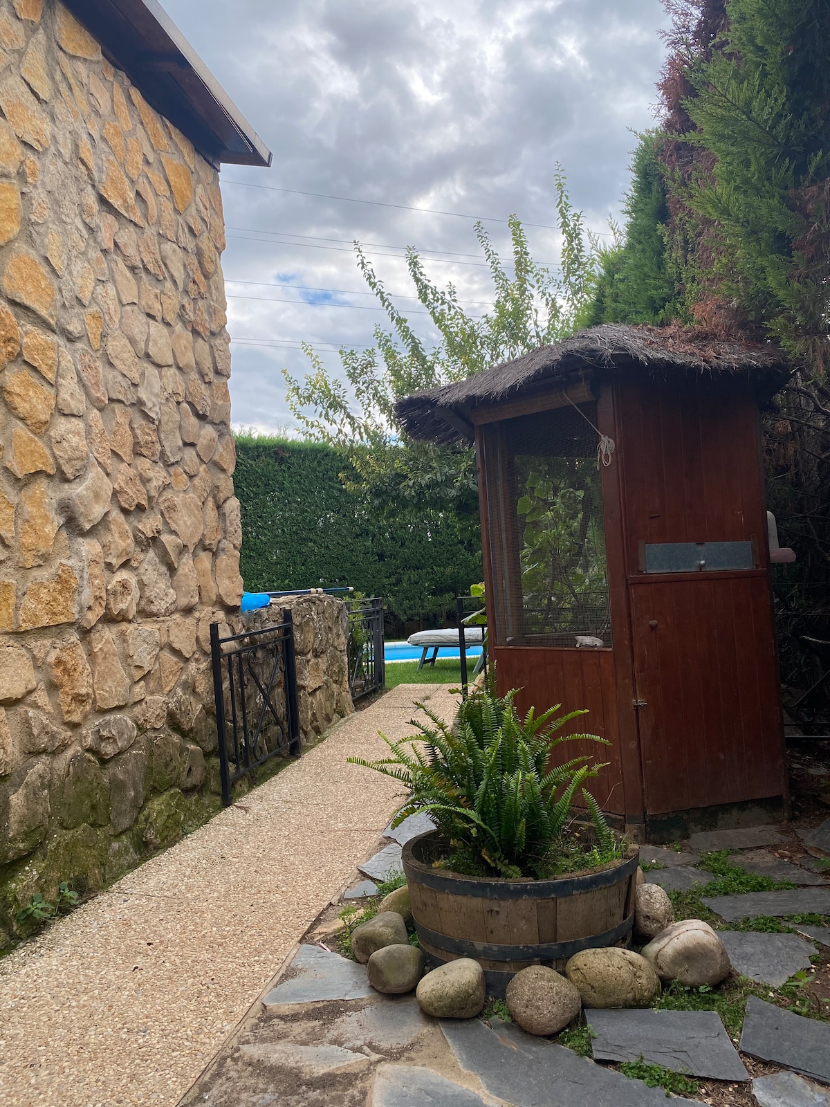 A pathway is lined with natural stone, leading past a wooden structure with thatched roofing. Lush greenery and a round planter filled with plants can be seen, alongside smooth pebbles. In the background, a glimpse of a turquoise pool is visible, framed by hedges.