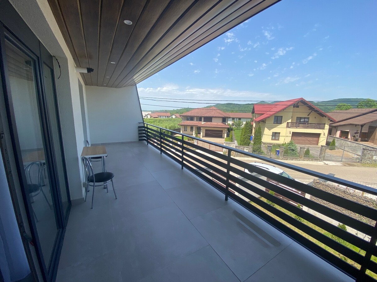 A generous balcony features modern railing and a table with a chair, providing an open space for relaxation. Scenic views of surrounding greenery and rooftops are visible, under a clear blue sky accentuated by soft clouds.