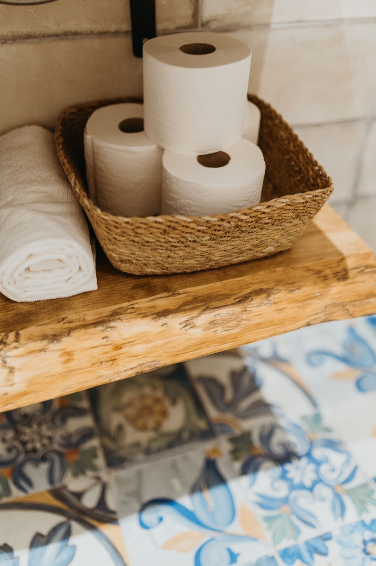A wooden shelf displays a woven basket containing several rolls of toilet paper alongside a neatly folded white towel. The floor features colorful patterned tiles, adding visual interest to the bathroom setting.
