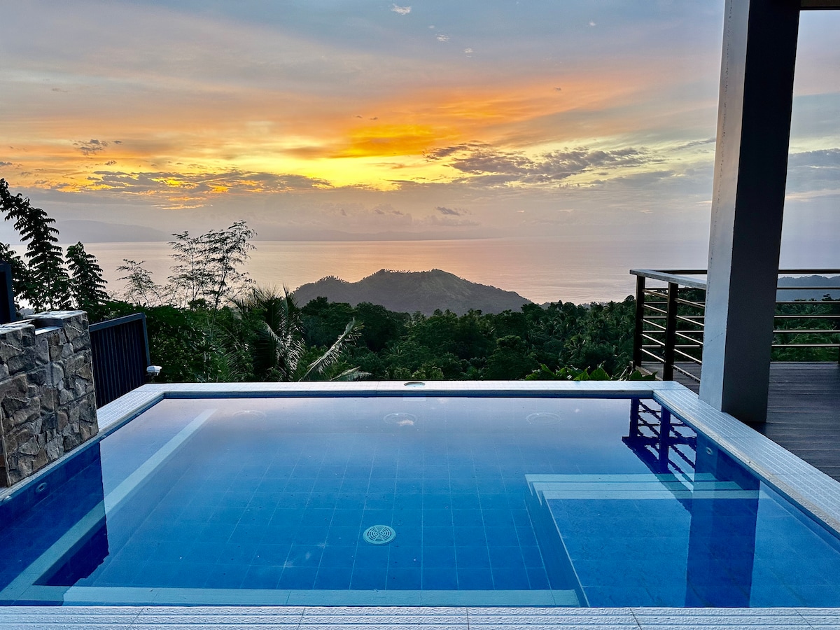An ocean-view infinity pool is framed by a stone wall, reflecting the vibrant colors of the sunset. Lush greenery surrounds the area, enhancing the serene ambiance. The horizon blends seamlessly with the sky, creating a tranquil backdrop for relaxation.