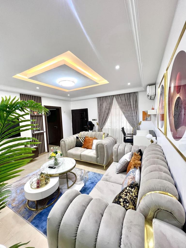A contemporary living area features a pair of plush sofas arranged around a round coffee table. A decorative plant adds greenery, while artwork adorns the walls. Soft lighting is provided by a tray ceiling, and large windows are dressed with light curtains.