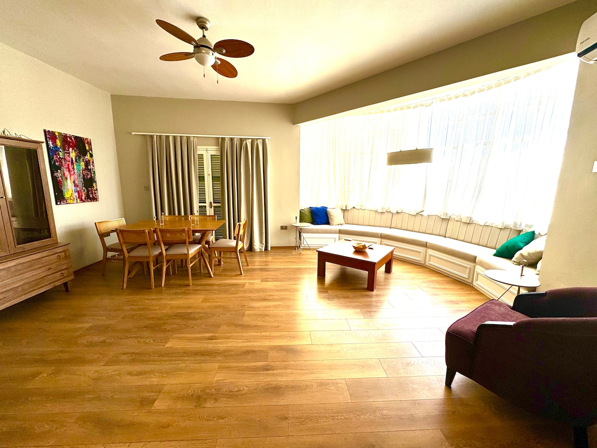 A spacious living area features large windows that allow natural light to fill the room. A wooden dining table with chairs sits opposite a comfortable seating area that includes a single armchair. Decorative cushions are placed on a built-in bench, enhancing relaxation.