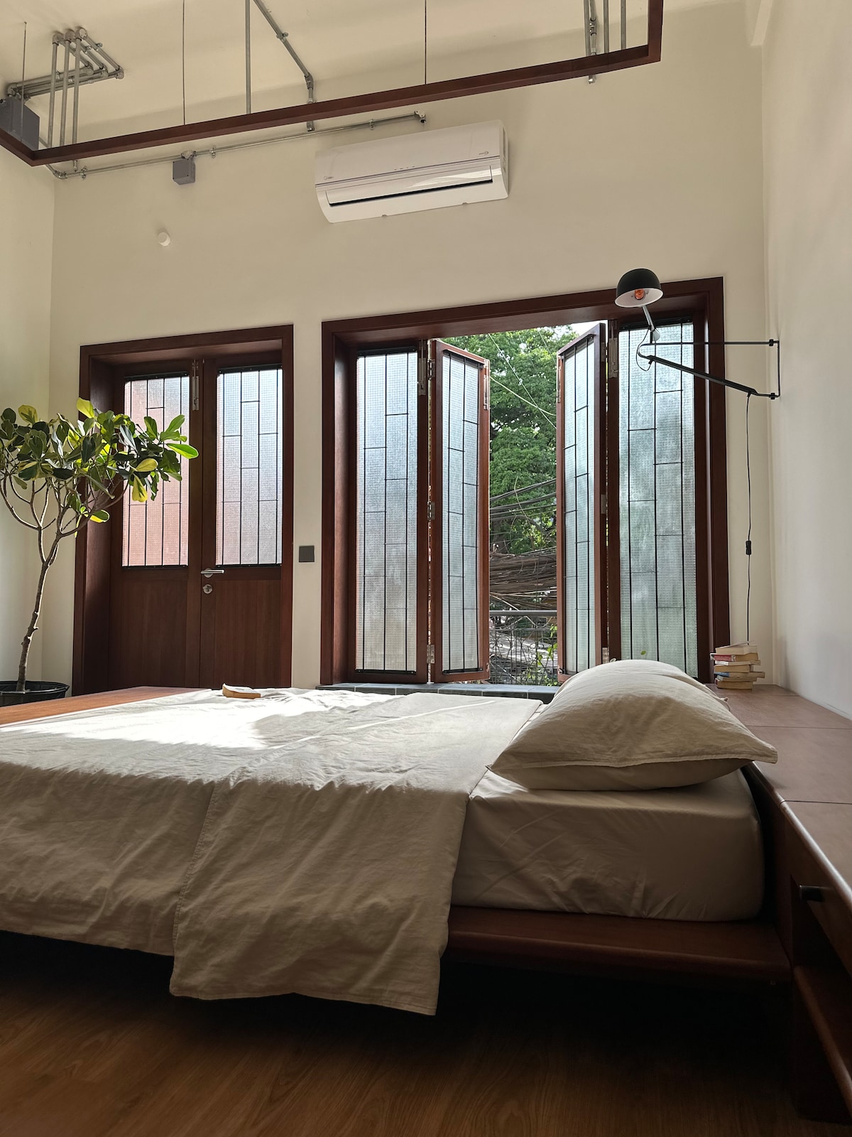 A bright bedroom features a spacious bed with a light-colored duvet and pillows, positioned near large windows that open to a leafy exterior. A small tree is located beside the bed, adding a touch of greenery. Natural light fills the room, enhancing its open feel.