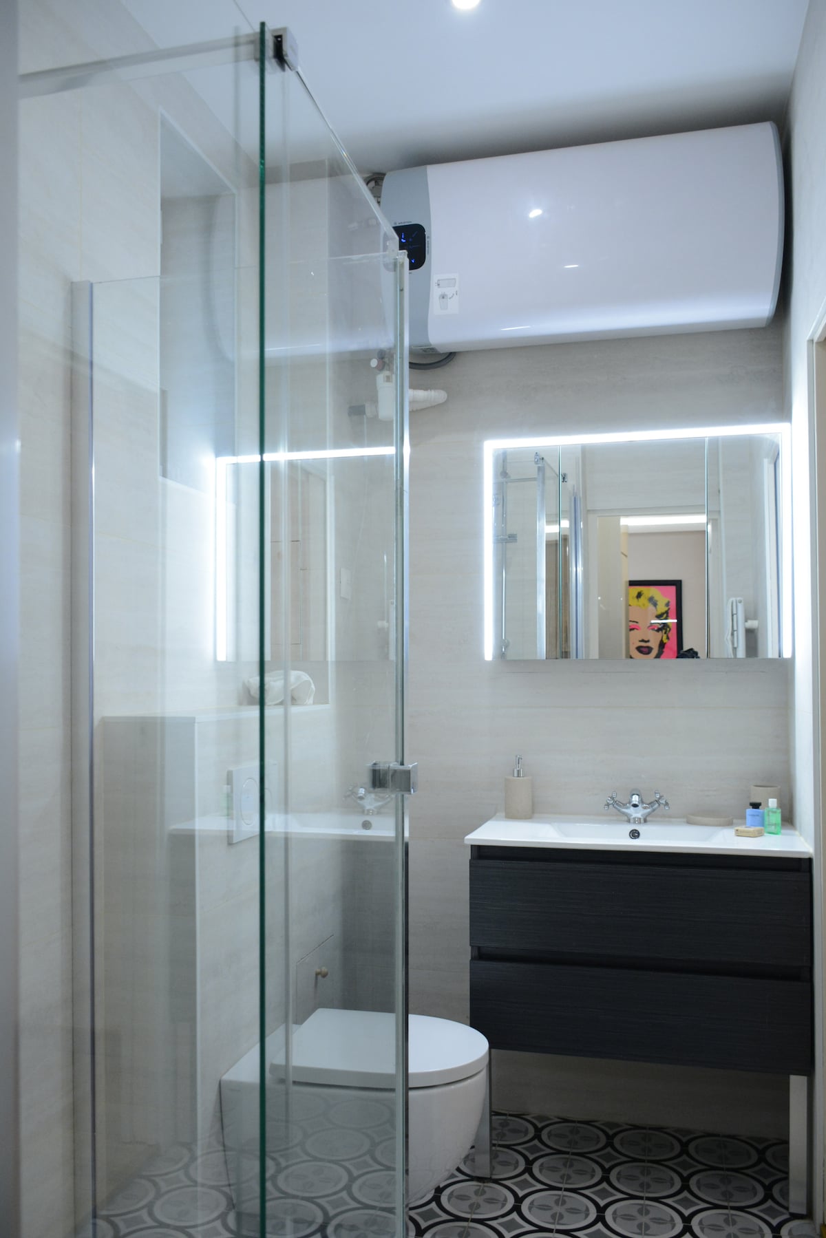 A modern bathroom features a walk-in shower enclosed by glass panels. The sleek sink is accompanied by a dark wooden cabinet with two drawers. Decorative tiles create a stylish contrast on the floor, and ample lighting enhances the space.