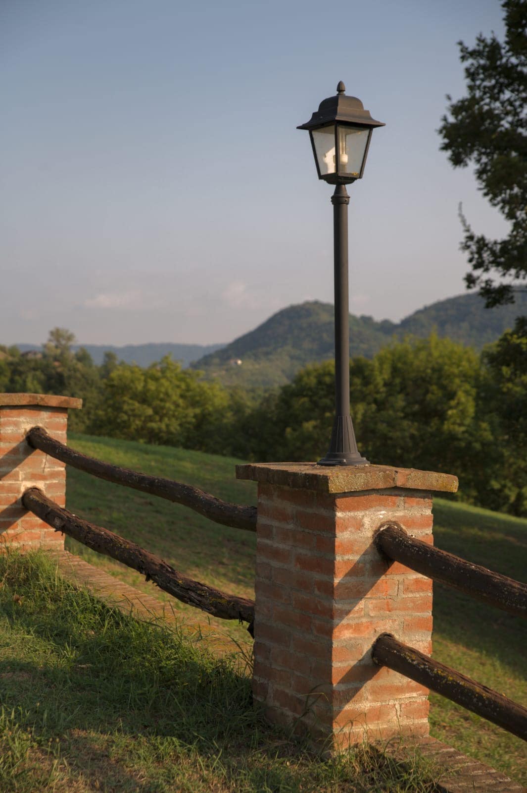 A classic street lamp is positioned on a brick post, providing soft illumination. The fixture stands near a rustic wooden railing, with rolling hills and lush greenery visible in the background, highlighting the natural beauty of the surrounding landscape.