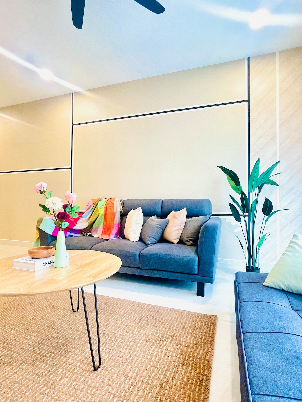 A comfortable living area features a grey sofa with decorative cushions, accompanied by a round coffee table. A vibrant throw blanket adds color to the space. Lush green plants provide a touch of nature, while a soft rug enhances the cozy atmosphere.