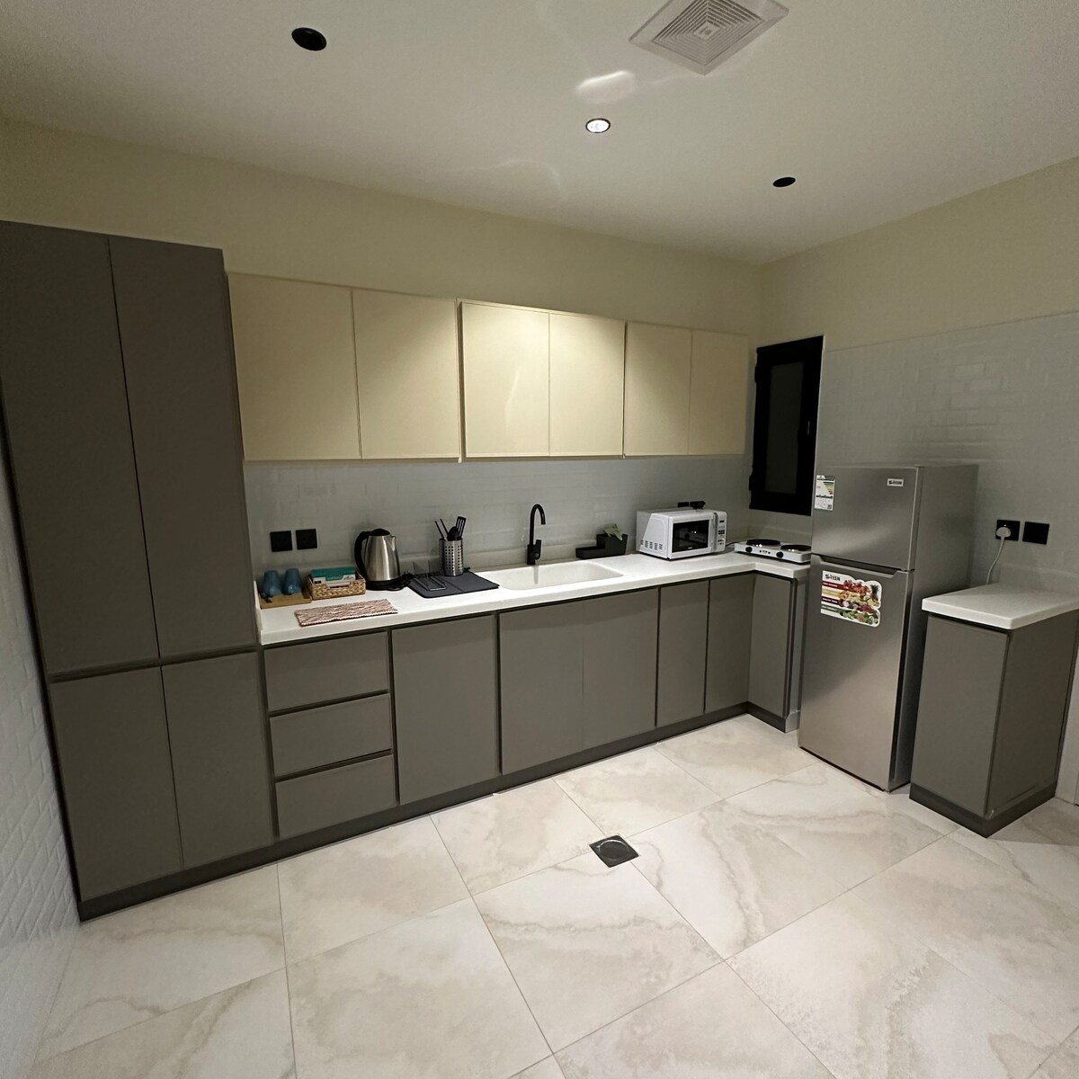 A modern kitchen is presented, featuring sleek cabinetry in neutral tones. Stainless steel appliances, including a refrigerator and microwave, are visible alongside a sink and kettle. Ample counter space is available, complemented by a light-colored tile floor that enhances the contemporary feel.