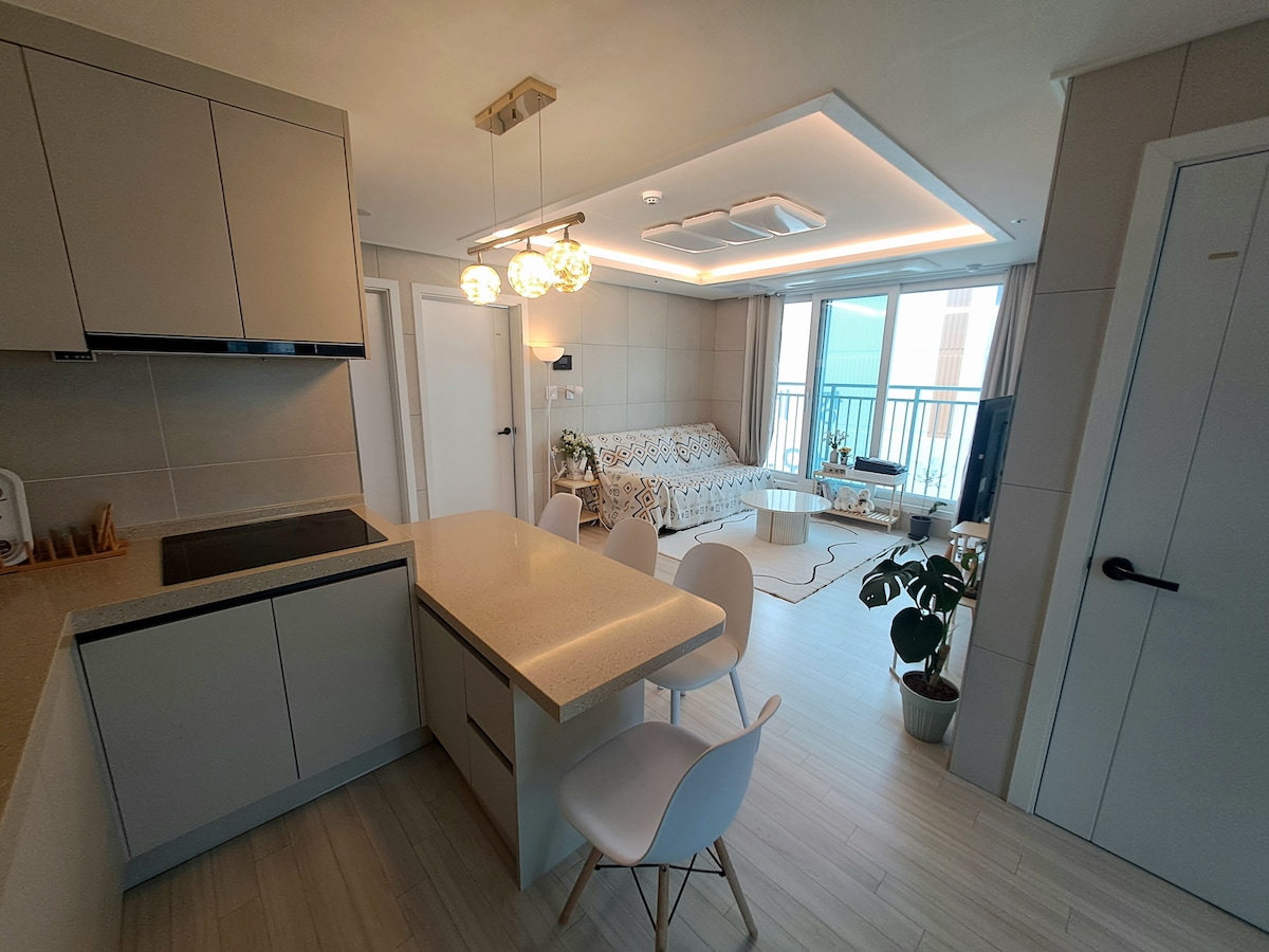 A modern, open kitchen area is present, featuring a spacious island with seating for four. A cozy living space is visible through large windows, complemented by plants and comfortable furnishings, while soft lighting enhances the neutral tones of the decor.