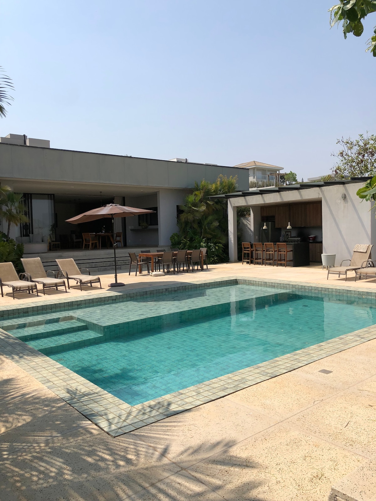 The outdoor area features a shimmering pool surrounded by lounge chairs, with an umbrella providing shade. A dining table is set nearby, complemented by a bar area. Lush greenery enhances the serene setting, ideal for relaxation and gatherings.