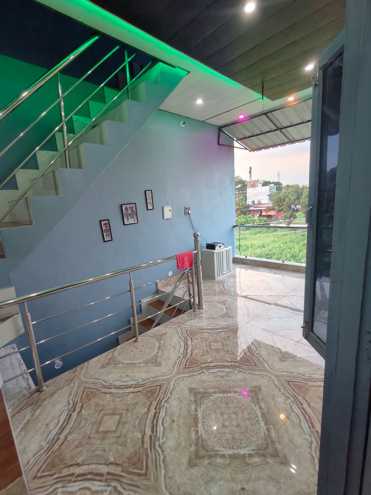 A spacious entrance area is illuminated by natural light streaming in through an open door. The staircase and railing are visible, along with decorative framed pictures on the blue wall. A modern tiled floor reflects the light, enhancing the airy feel of the space.