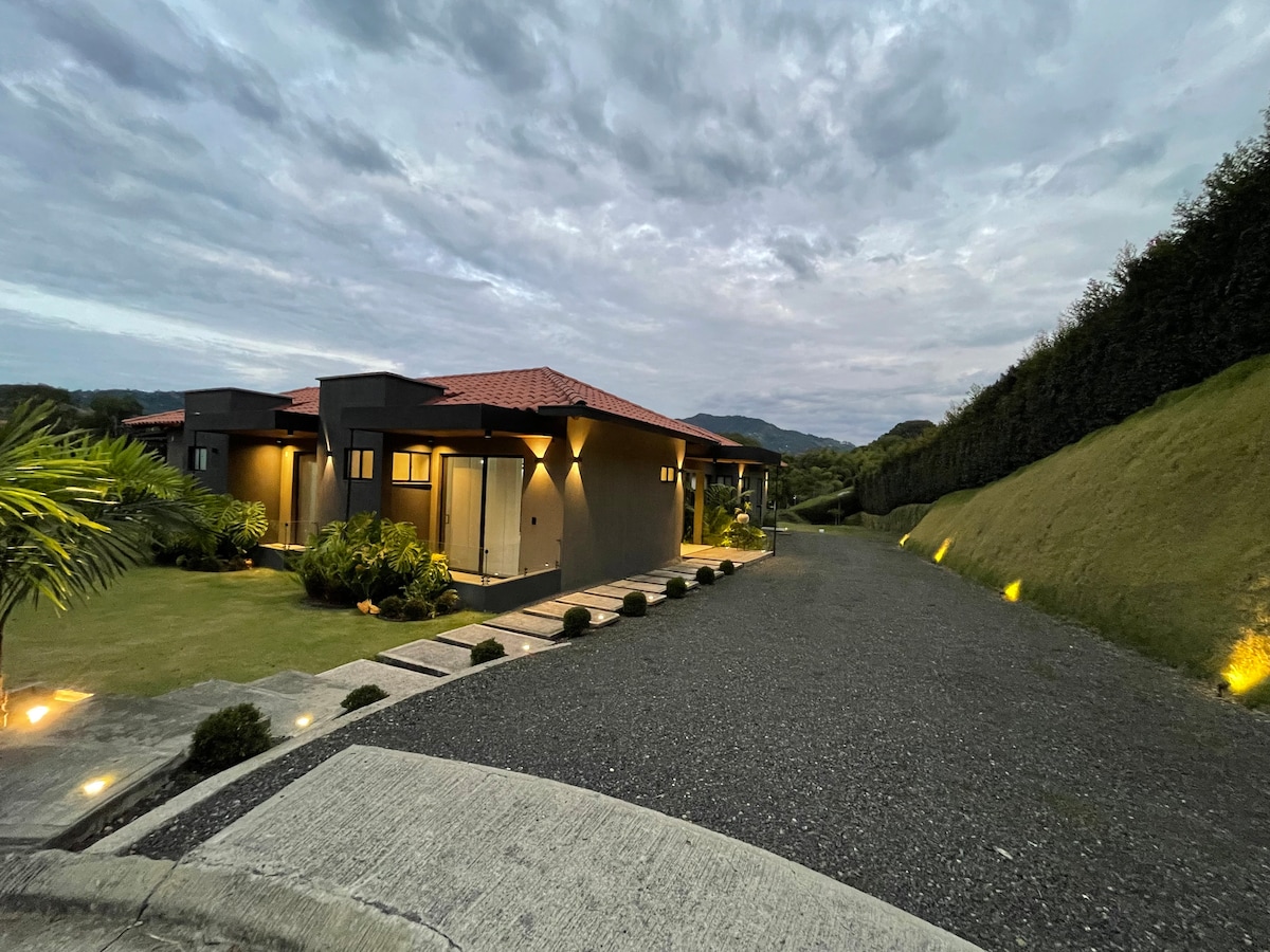 A modern country home is set against a backdrop of gently rolling hills, surrounded by lush greenery. The exterior features a red-tiled roof and large windows, while a well-defined pathway leads to the entrance, illuminated by soft outdoor lights.