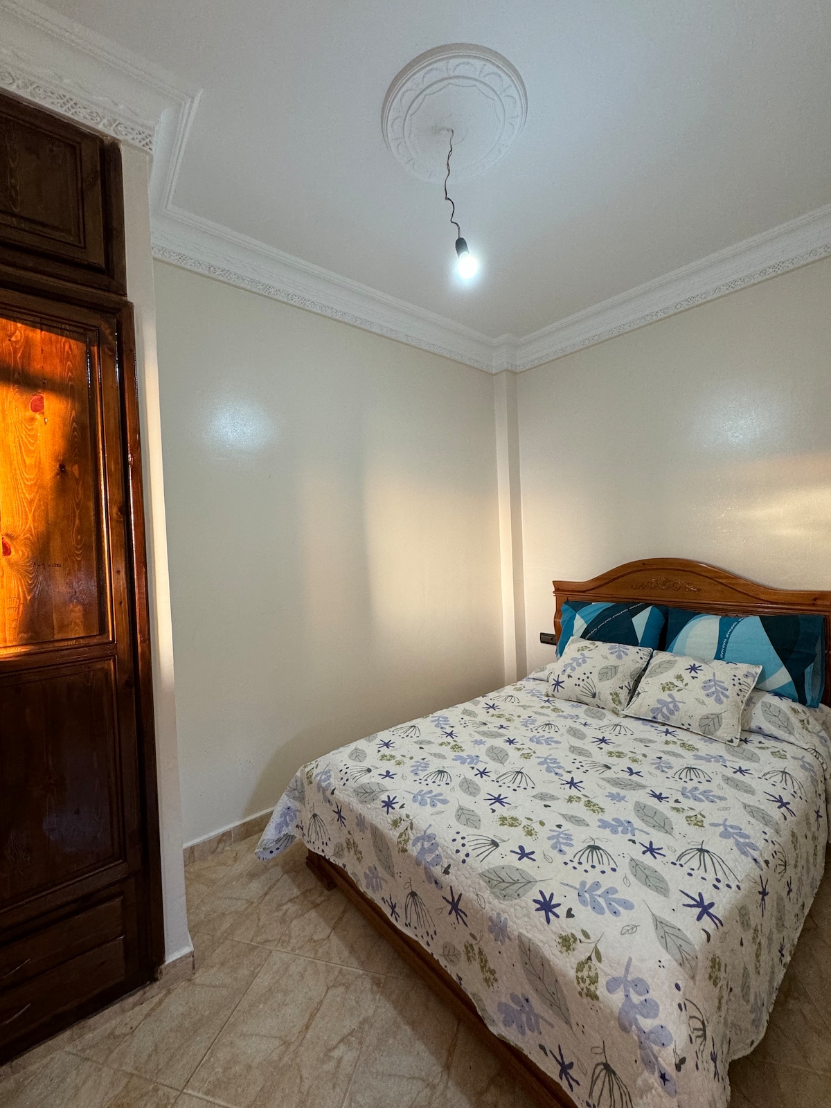 A well-lit bedroom features a spacious bed adorned with a floral-patterned blanket and multiple pillows. A wooden wardrobe stands against the wall, contributing to the simple decor. Soft natural light highlights the neutral-colored walls, creating a serene atmosphere.