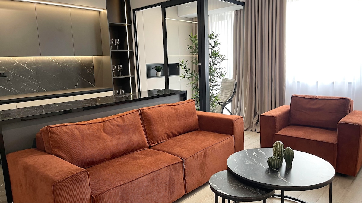 A modern living area features a plush sectional sofa and two armchairs in warm tones. A round coffee table sits in front, adorned with decorative items. Natural light filters through sheer curtains, while a sleek kitchenette is visible in the background.