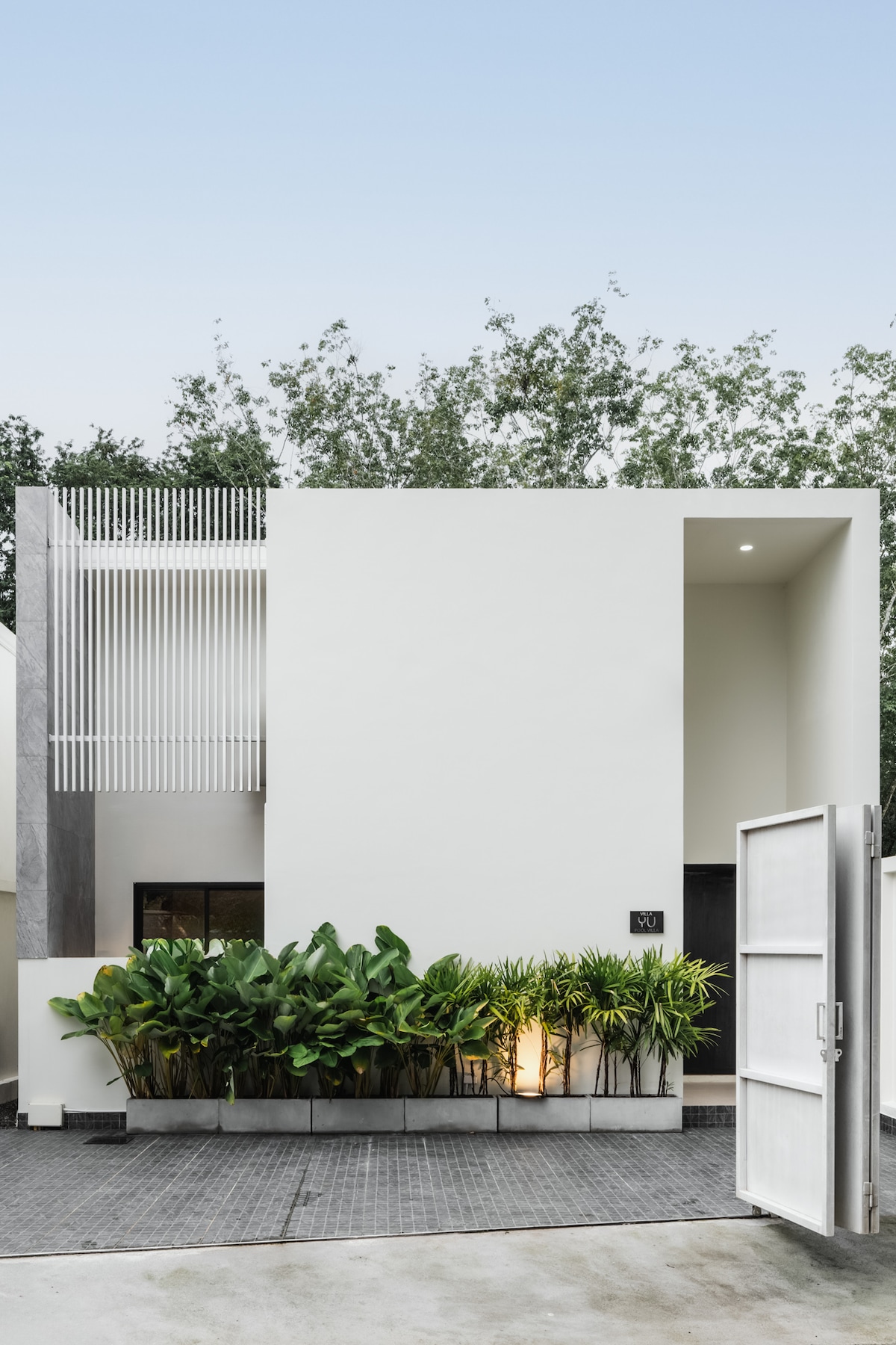The exterior of a contemporary villa is showcased, featuring a minimalist design with clean lines and large windows. Lush greenery and well-maintained plants are positioned along the entrance, enhancing the natural ambiance. A welcoming open door invites guests to explore the tranquil surroundings.