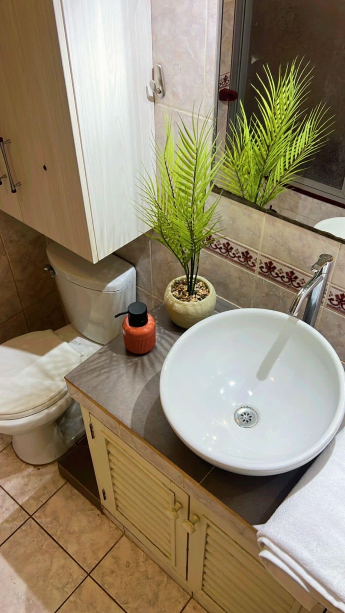 The bathroom features a sleek round sink atop a counter, complemented by a small potted plant. A toilet is positioned nearby, and a cupboard above provides storage. Neutral tones and tiled surfaces contribute to a clean and organized appearance.