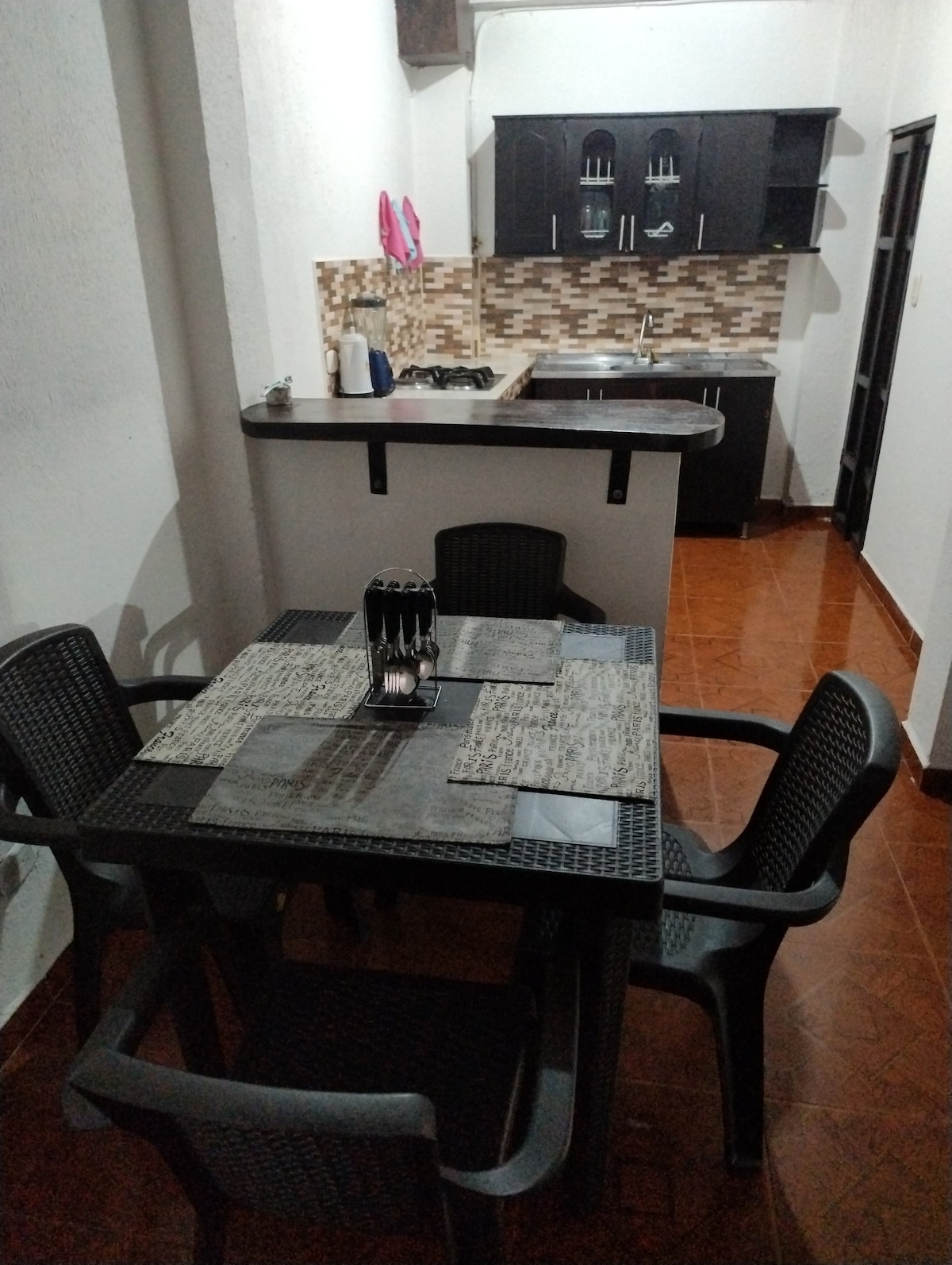 A dining area features a dark rectangular table accompanied by four textured chairs, set with placemats and cutlery. The open kitchen is visible in the background, showcasing modern cabinetry and a tiled backsplash.