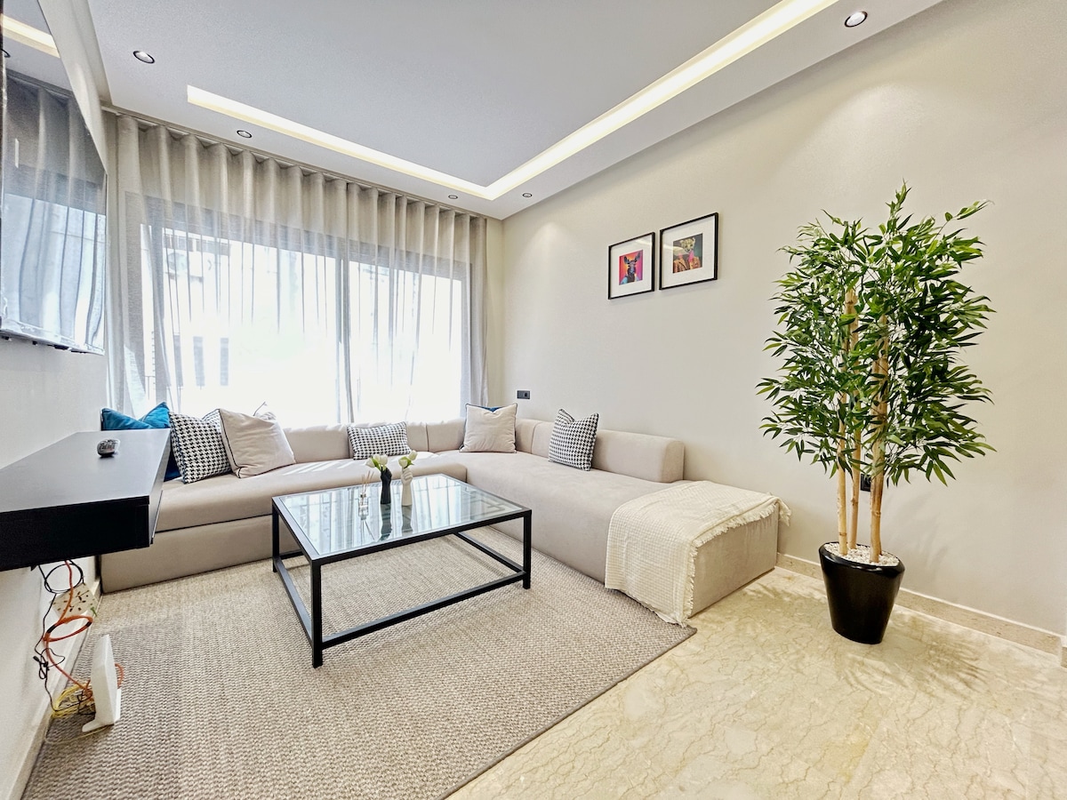 A modern living area is presented with a spacious sectional sofa adorned with patterned cushions. A glass coffee table sits atop a textured rug, while a decorative plant adds a touch of greenery. Natural light floods the room through sheer curtains framing a large window.