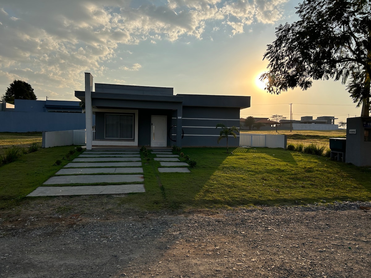 A modern house is shown in the foreground, framed by a manicured lawn and stone pathways. The sun sets in the background, casting a warm glow over the scene. Clear skies and open space contribute to a serene outdoor environment.