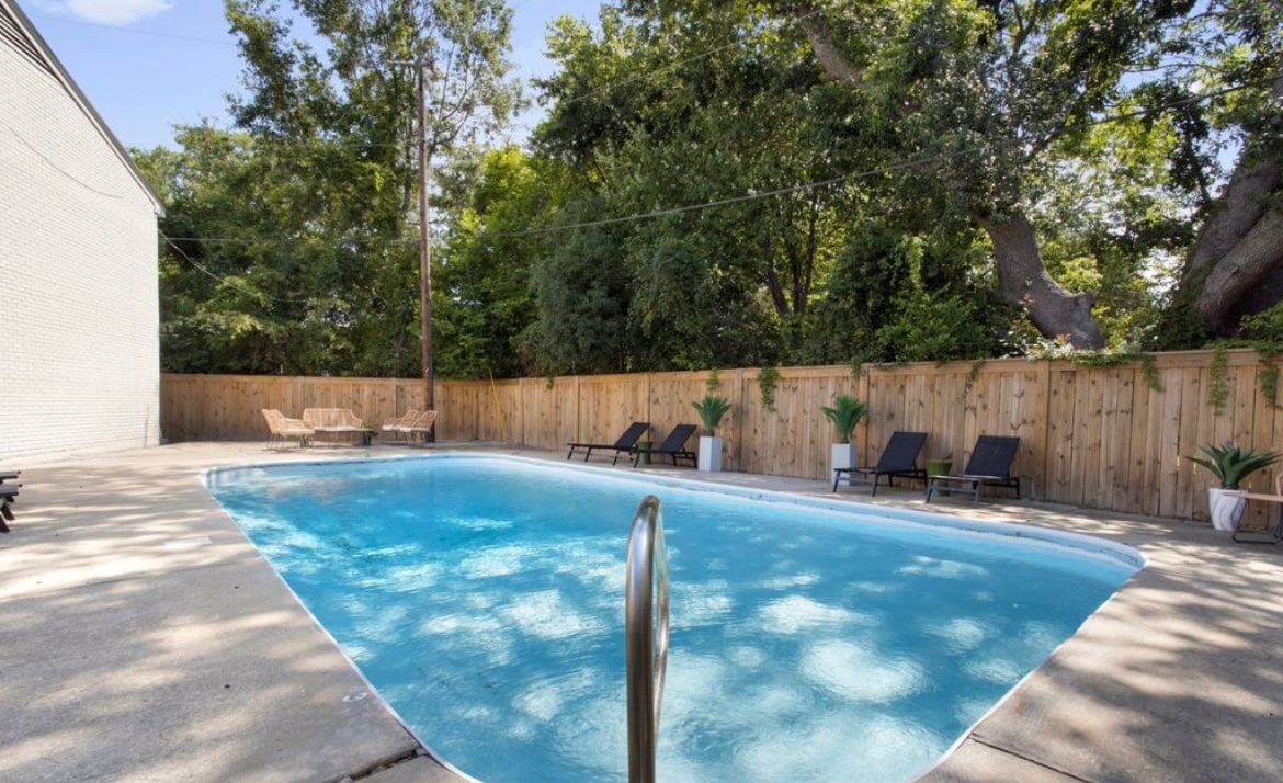 A private pool area is surrounded by a wooden fence, offering a serene retreat. Several lounge chairs are positioned around the pool, with lush greenery visible in the background. The clear water of the pool reflects the shade of trees above, creating a tranquil atmosphere.