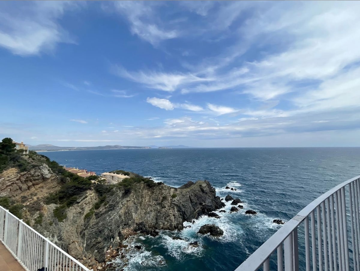 The image captures a sweeping view of the rocky coastline meeting the sea under a partly cloudy sky. Waves can be seen crashing against the rocks, while the horizon extends into the distance, showcasing a tranquil blue sea.