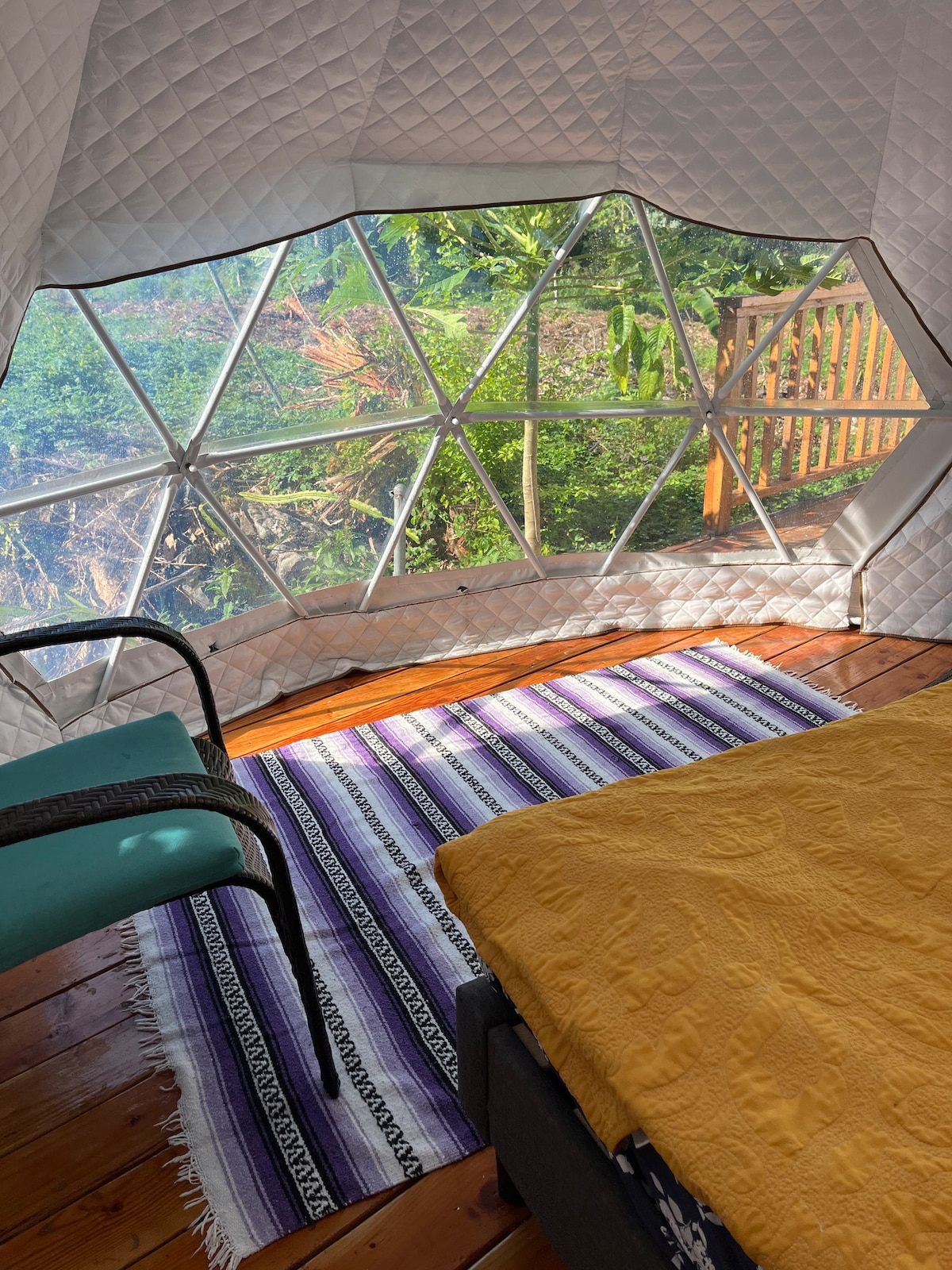 A cozy interior features a spacious bed covered with a yellow quilt, complemented by a patterned area rug. Large geodesic windows allow natural light to fill the space, providing views of the surrounding greenery and inviting the beauty of nature inside.