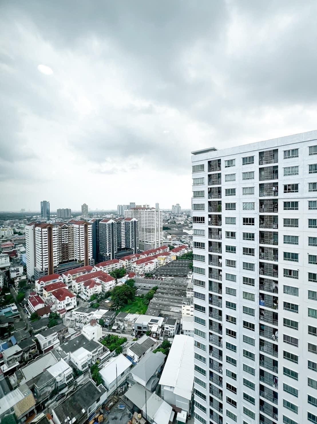 A cityscape is captured from a high vantage point, showcasing a mix of modern and traditional architecture. Rows of residential buildings lay amidst urban greenery, with distant skyscrapers lining the horizon under a cloudy sky.