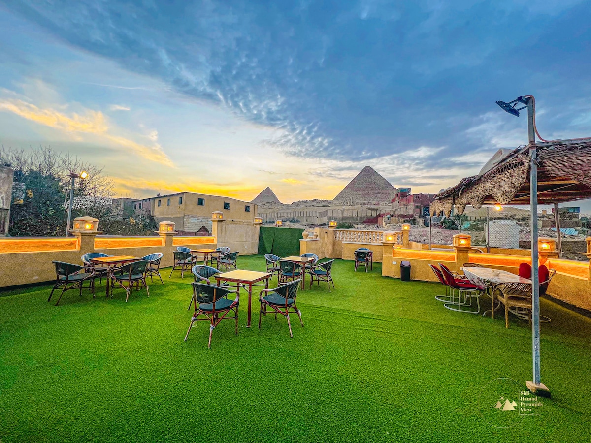 A rooftop terrace is presented, featuring green artificial turf and various seating arrangements. Tables and chairs are scattered throughout, alongside shaded lounge areas. The backdrop includes pyramids and a colorful sunset sky, creating a serene environment for gatherings.