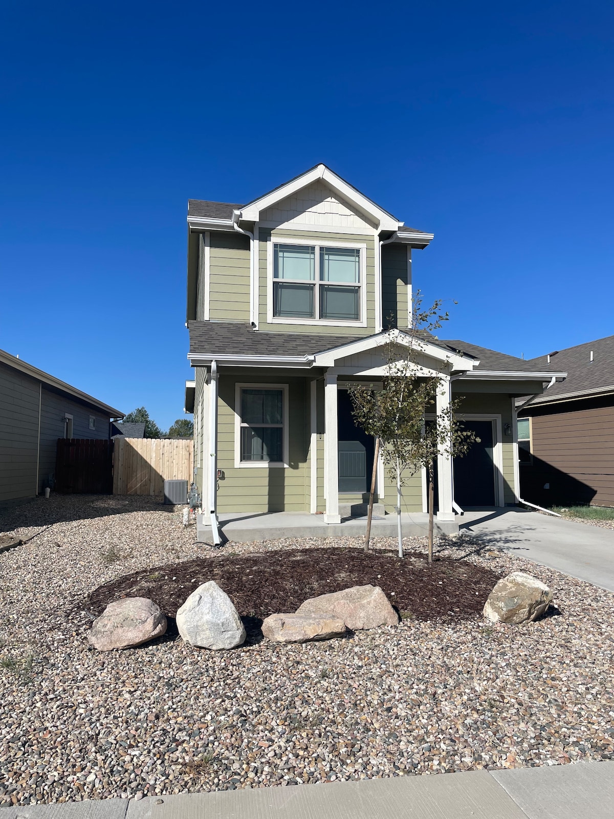 A modern two-story house features a welcoming entrance with white pillars and a small tree planted in the landscaped front yard. The exterior is painted in a soft green tone, complemented by large windows that invite natural light. A gravel pathway leads to the home.