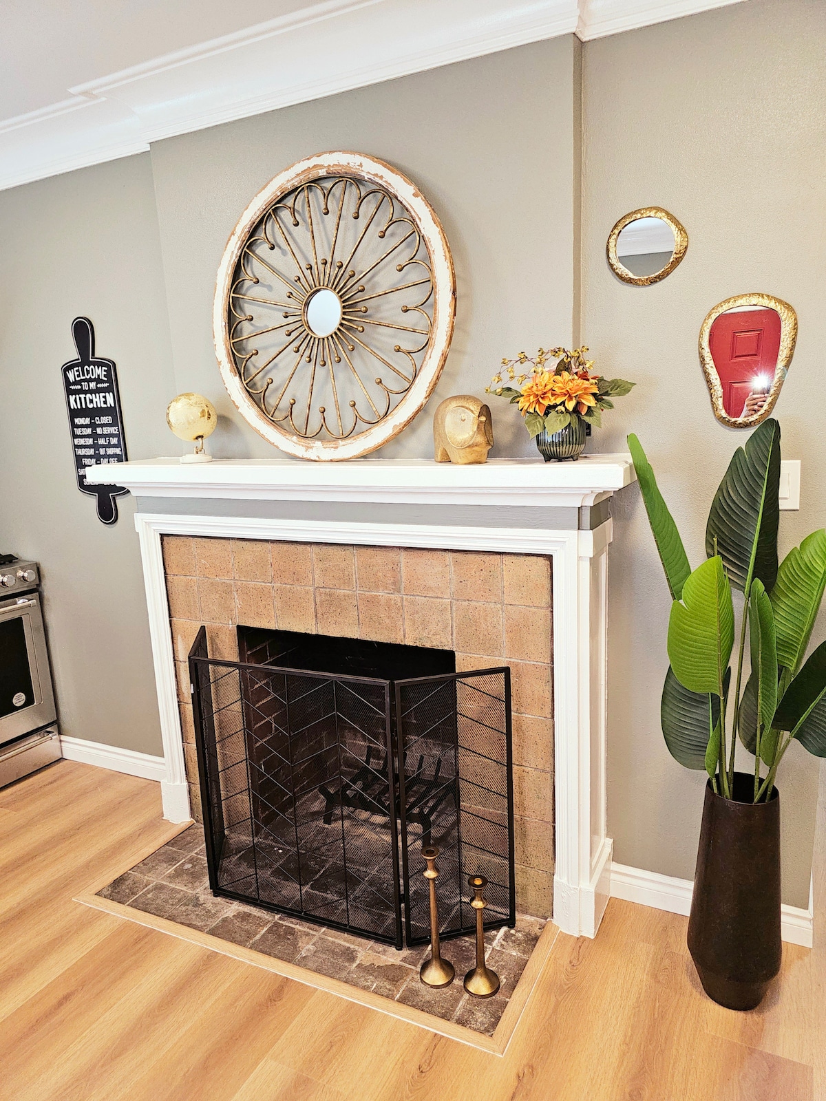A cozy fireplace is framed by a decorative mantel, featuring a round mirror and wall art. A tall potted plant stands beside the hearth, and small decorative items are displayed along the mantel. The warm wall color complements the wooden flooring.