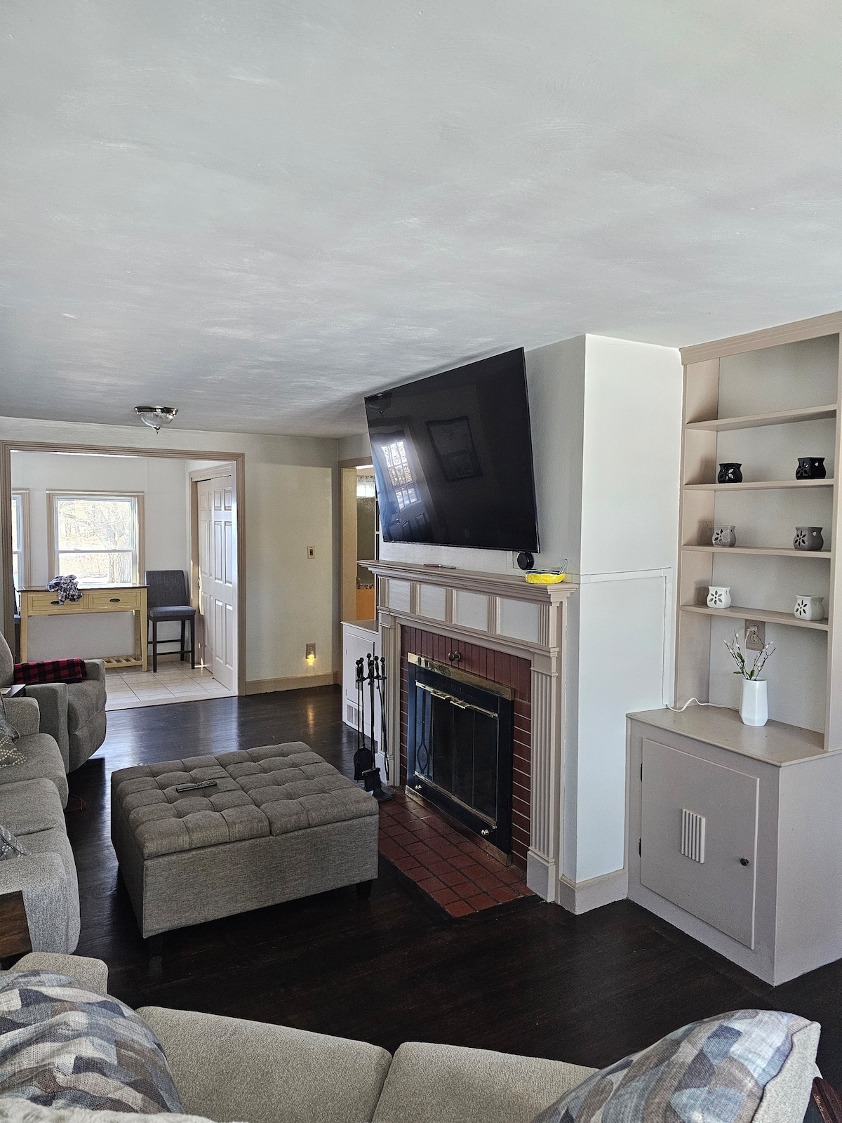 A spacious living area is featured, highlighted by a large flat-screen television mounted above a classic fireplace. Comfortable seating includes a sectional sofa and an ottoman, positioned around the fireplace. A shelving unit and a doorway leading to another room are visible.