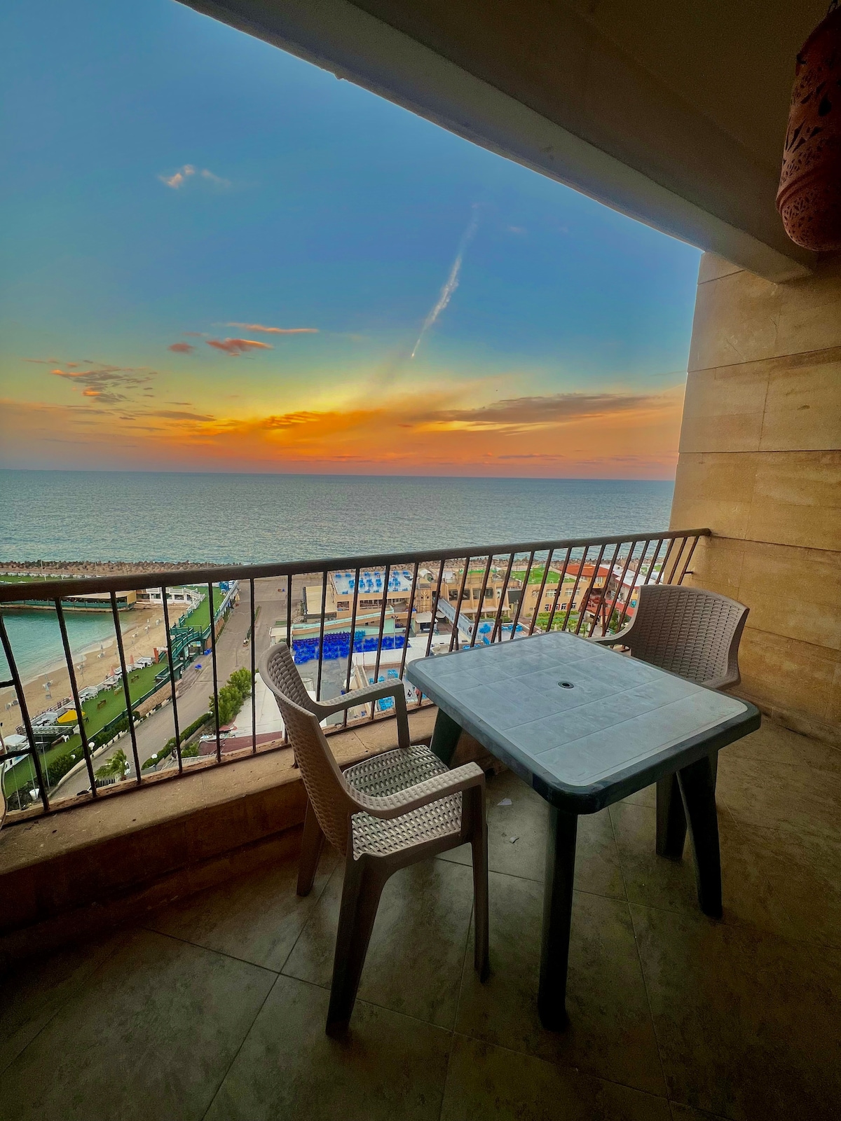A balcony overlooks the stunning sea view at sunset, featuring a small table with two chairs. The horizon is painted with warm hues, and the gentle waves of the ocean create a serene atmosphere, inviting relaxation and enjoyment of the coastal scenery.