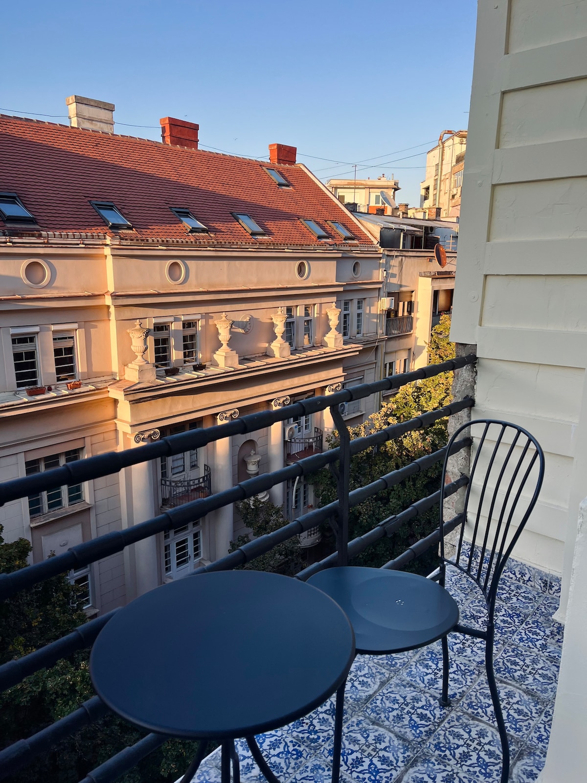 A small balcony features a round table and two metal chairs, set against a backdrop of classic architecture. The intricate tile flooring adds a decorative touch, while the warm light of the setting sun highlights the building's details across the street.