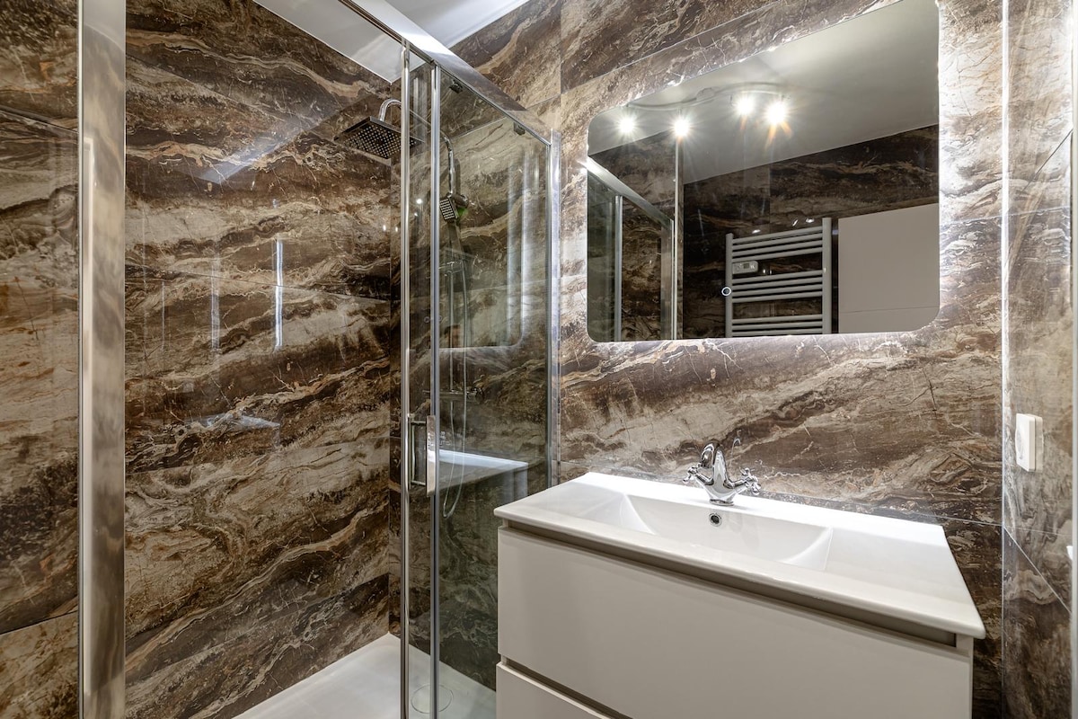 The bathroom features a glass shower enclosure and a modern sink with a sleek countertop. Elegant stone wall tiles create a sophisticated backdrop, complemented by illuminated mirrors. A heated towel rail is positioned nearby, enhancing the functional space.