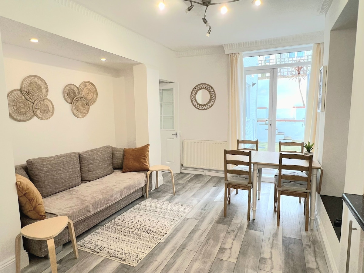 A modern living area features a gray sofa accented with decorative pillows, paired with a wooden table and four wooden chairs. Natural light floods the space through large glass doors leading to the courtyard, complemented by minimalist wall decor.